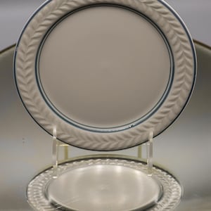 Op de afbeelding: Een witte keramische plaat met een lichtblauwe bladerenpatroonrand en een blauwe accentlijn. De plaat wordt gepresenteerd op een transparante standaard, met de weerspiegeling eronder zichtbaar.