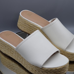 Può includere: Un paio di sandali bianchi a fascia con un cinturino largo sulla parte superiore e una spessa suola platform rivestita in materiale di juta intrecciata color marrone naturale. Il plantare è color marrone chiaro con la scritta True Soul stampata.