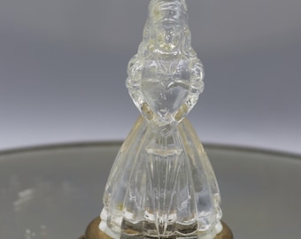 Puede incluir: Un frasco de perfume de vidrio transparente con un diseño en forma de figura. El frasco tiene una tapa y una base de metal dorado. La figura parece estar sosteniendo un corazón. El frasco está sobre una superficie reflectante.