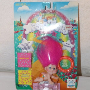 Könnte beinhalten: Eine rosa und blaue Magic Troll Puppe mit einer regenbogenfarbenen Perücke. Die Puppe ist in einer Plastik-Blisterverpackung mit einem Regenbogenhintergrund und den Worten "Magic Trolls" in großen Buchstaben verpackt. Die Puppe ist voll beweglich und wird mit einem magischen Glücksarmband geliefert.
