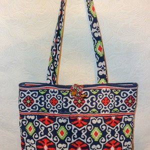 Puede incluir: Un bolso tote acolchado azul y blanco con un patrón geométrico rojo, verde y blanco. El bolso tiene un cierre de botón de madera marrón y dos correas largas.