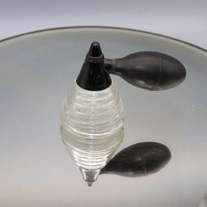 May include: A clear glass perfume bottle with a black plastic pump and a black rubber bulb. The bottle has a ribbed design and is sitting on a reflective surface.
