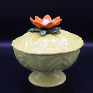 May include: A yellow ceramic sugar bowl with a lid and a flower on top. The bowl has a fluted design and a pedestal base.