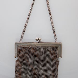 May include: An antique silver chainmail purse with a rectangular frame and a chain handle. The purse features a detailed woven chainmail design in shades of silver and brown, with a clasp closure.
