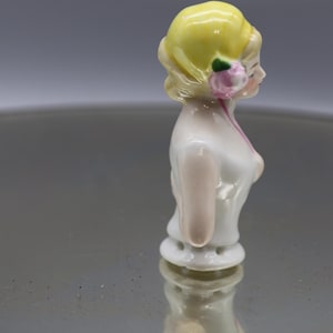 May include: A porcelain figurine of a woman, featuring a yellow updo, a pink flower, and a white dress with pink accents. The figurine is standing upright, with a detailed face and delicate features. The overall aesthetic is vintage.