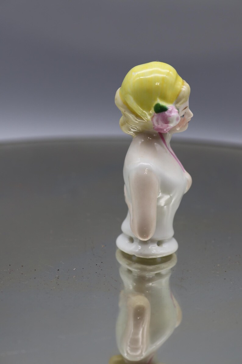 May include: A porcelain figurine of a woman, featuring a yellow updo, a pink flower, and a white dress with pink accents. The figurine is standing upright, with a detailed face and delicate features. The overall aesthetic is vintage.