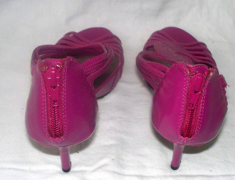 May include: A pair of bright pink high-heeled sandals with a zipper detail on the back. The sandals have a strappy design and are made of a shiny material.