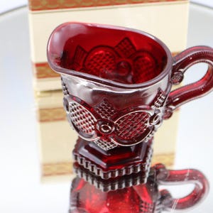 Op de afbeelding: Een kleine, robijnrode glazen creamer met een handvat en sierlijke details. De creamer heeft een textuurpatroon en staat op een reflecterend oppervlak. Een decoratieve doos staat op de achtergrond.