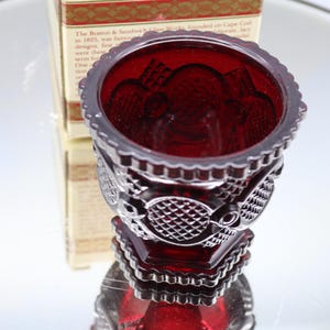 Op de afbeelding: Een robijnrode glazen votiefkaarsenhouder met ingewikkelde reliëfpatronen. De houder heeft een geschulpte rand en een geometrische basis. De doos erachter heeft de tekst "The Boston & Sandwich Glass Works founded on Cape Cod".