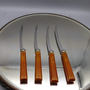May include: Four cheese knives with curved stainless steel blades and amber-colored handles are arranged on a round mirror. The knives are evenly spaced, reflecting in the mirror's surface. The mirror has a decorative, ornate edge.