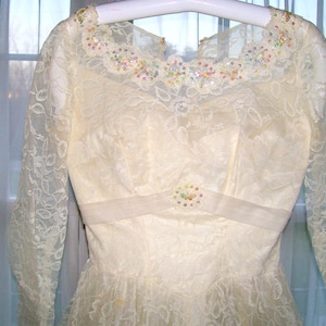 Puede incluir: Un vestido de novia vintage de encaje marfil con un escote de cuentas y una faja de satén. El vestido tiene mangas largas y una falda completa.