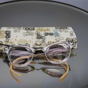 May include: Vintage cat-eye eyeglasses with a silver frame and light brown temples. The glasses rest in front of a patterned case with a cream background and black and gold designs. The lenses are clear.