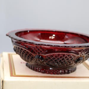 May include: A ruby red glass bowl with a decorative, textured pattern. The bowl has a scalloped rim and sits on a small, round base. The bowl is likely vintage and is displayed on a white box.