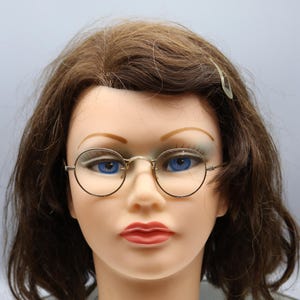 May include: A mannequin head with brown hair and blue eyes wearing round, gold-rimmed eyeglasses. The mannequin has red lipstick and a white collar with the text "Patty" on it. A clear hair clip is in the hair.
