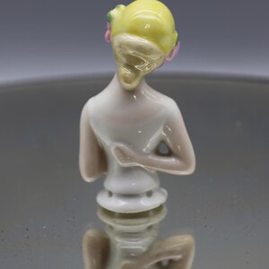 May include: A porcelain figurine of a woman's torso, viewed from the back. The figure has a yellow head with pink and green accents. The body is white with subtle pink shading. The figurine is a decorative item.