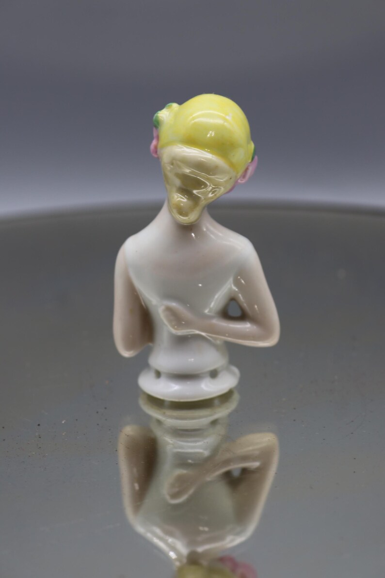 May include: A porcelain figurine of a woman's torso, viewed from the back. The figure has a yellow head with pink and green accents. The body is white with subtle pink shading. The figurine is a decorative item.