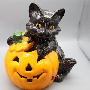 Könnte beinhalten: Keramik-Halloween-Dekoration mit einer schwarzen Katze auf einem lächelnden orangefarbenen Kürbis. Die Katze hat gelbe Augen, der Kürbis hat ausgeschnittene Augen, Nase und Mund. Eine festliche Dekoration.