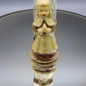 May include: A vintage figurine of a girl in a dress, hands clasped in prayer, wrapped in clear plastic. The figurine has blonde hair, blue eyes, and a red mouth. The dress is light yellow and brown.