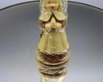May include: A vintage figurine of a girl in a dress, hands clasped in prayer, wrapped in clear plastic. The figurine has blonde hair, blue eyes, and a red mouth. The dress is light yellow and brown.