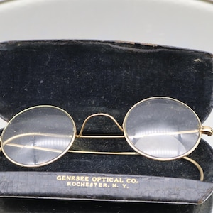 May include: Vintage round eyeglasses with a gold-tone frame, resting in a black case. The case is open, revealing the glasses and the text "GENESEE OPTICAL CO. ROCHESTER, N. Y." The glasses have clear lenses and a delicate bridge.