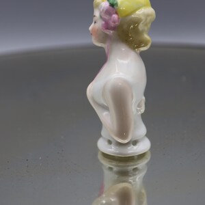 May include: A vintage porcelain figurine of a woman's bust, featuring a yellow cap adorned with a pink flower and green leaves. The figurine has a white bodice and is reflected on a shiny surface.