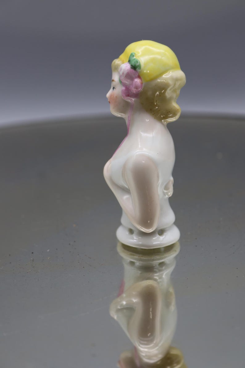May include: A vintage porcelain figurine of a woman's bust, featuring a yellow cap adorned with a pink flower and green leaves. The figurine has a white bodice and is reflected on a shiny surface.