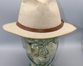 May include: A beige fedora-style hat with a brown leather band. The hat has a structured crown and a wide brim. The hat is displayed on a glass head form against a gray background. The hat appears to be made of a felt-like material.