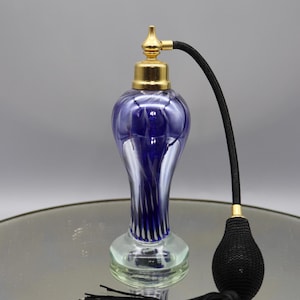 May include: An antique perfume bottle with a blue and white swirled glass body, a gold-colored metal top, and a black bulb atomizer with a tassel. The bottle has a clear glass base and sits on a reflective surface.