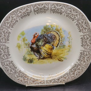 Puede incluir: Plato ovalado para servir con una ilustración de pavo. El plato de cerámica blanca tiene un patrón floral marrón alrededor del borde y las asas. El pavo está representado en un campo con una valla y árboles al fondo.