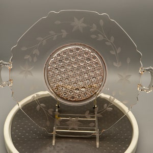 May include: A vintage pink glass serving dish with etched floral designs and a raised, textured center. The dish has two handles and a scalloped edge.