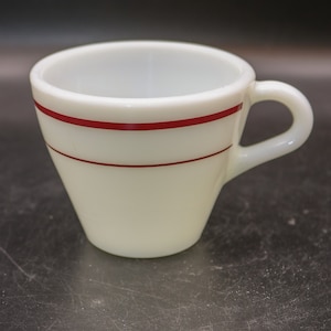 Könnte beinhalten: Eine weiße Keramiktasse mit zwei roten Streifen um den oberen Rand. Die Tasse hat einen abgerundeten Boden und einen Henkel.