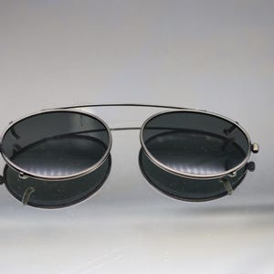 Puede incluir: Gafas de sol con montura plateada y lentes oscuras. Las gafas tienen un doble puente y una forma redonda clásica. Las lentes son oscuras, proporcionando sombra. Las gafas se reflejan en la superficie de abajo.