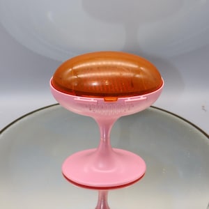 Könnte beinhalten: Eine rosa und orangefarbene Plastik-Schmuckschale mit einem Kuppeldeckel. Die Schale hat einen Sockel und steht auf einer spiegelnden Oberfläche.