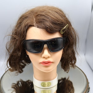 May include: A black pair of sunglasses with a dark lens. The sunglasses are on a mannequin head with brown hair. The mannequin is wearing a white collar with a gold label that says "Betty  Fidan" on it.