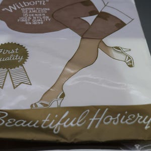 Puede incluir: Un paquete de medias vintage con un dibujo de piernas con medias con costuras y tacones altos. El paquete es blanco con detalles en marr&oacute;n y dorado, y se ve el texto "Beautiful Hosiery".