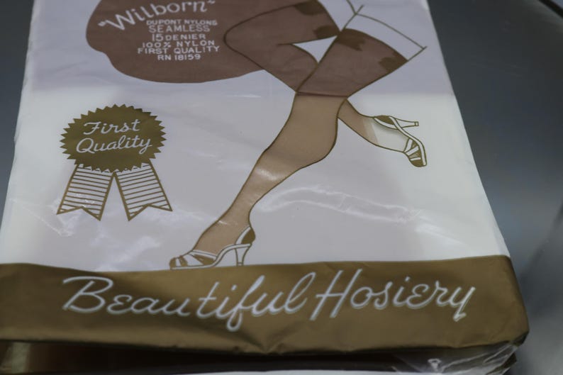 Puede incluir: Un paquete de medias vintage con un dibujo de piernas con medias con costuras y tacones altos. El paquete es blanco con detalles en marr&oacute;n y dorado, y se ve el texto "Beautiful Hosiery".