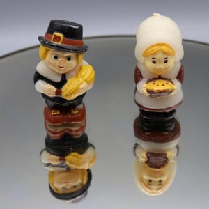 May include: Two small Thanksgiving figurines. One is a pilgrim man holding corn, wearing a black hat with a gold buckle, a black coat, and brown pants. The other is a pilgrim woman holding a pie, wearing a white bonnet and a brown dress.