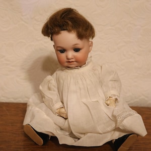 May include: A porcelain doll with brown hair wearing a white dress and black shoes. The doll is sitting on a wooden surface.