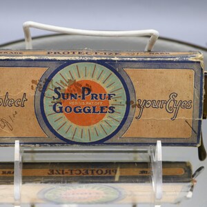 Op de afbeelding: Vintage Sun-Pruf Goggles doos met een witte handgreep. De doos is bruin met blauwe en oranje accenten en heeft de tekst "Sun-Pruf Goggles" en "Protect your Eyes". De doos wordt op een transparante standaard getoond.