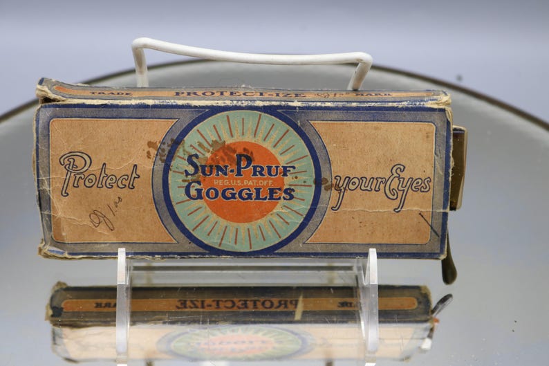Op de afbeelding: Vintage Sun-Pruf Goggles doos met een witte handgreep. De doos is bruin met blauwe en oranje accenten en heeft de tekst "Sun-Pruf Goggles" en "Protect your Eyes". De doos wordt op een transparante standaard getoond.