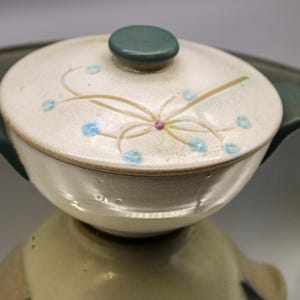 May include: A white ceramic serving dish with a teal lid and a floral design. The dish has a blue and pink floral pattern with a teal rim.