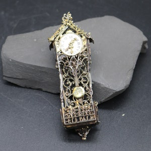 May include: A gold and black miniature grandfather clock with a white face and black Roman numeral hour markers. The clock has a decorative, ornate design.