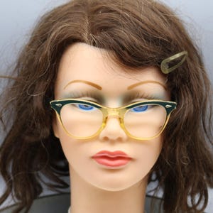 May include: Vintage cat-eye eyeglasses with a yellow and green frame. The glasses have a classic design, with a slightly upswept shape. The frame is a combination of yellow and green, with a clear bridge and temples. The lenses are clear.
