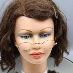 May include: A mannequin head with brown hair and blue eyes wearing gold-rimmed oval eyeglasses. The mannequin has a fair complexion and is wearing a white collar with the text "Pretty" on it.