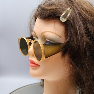 Op de afbeelding: Zonnebril in vintage-stijl met een lichtbruin montuur en donkere glazen. De zonnebril heeft een ronde vorm en een dik montuur. Er is ook een goudkleurige haarclip zichtbaar. De zonnebril wordt getoond op een mannequinhoofd.