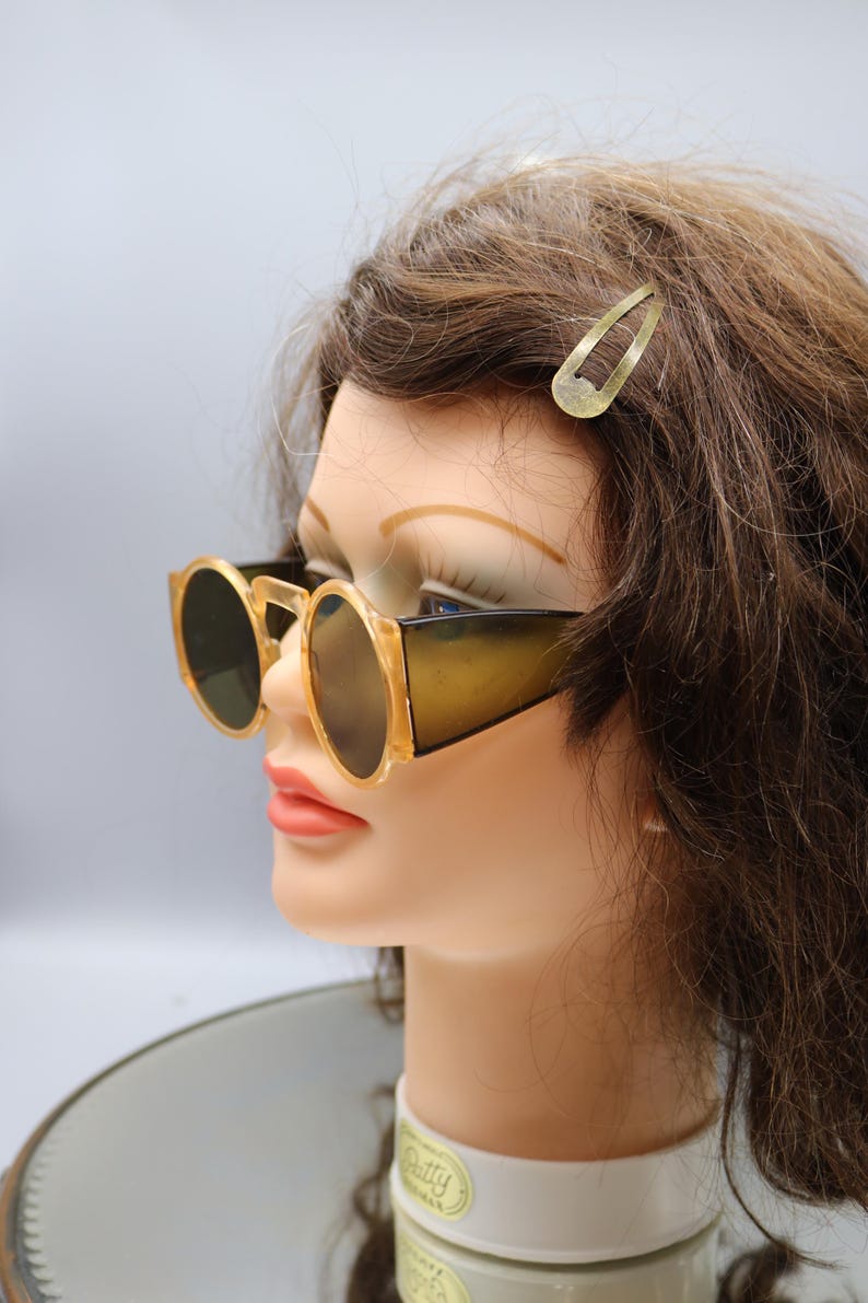 Op de afbeelding: Zonnebril in vintage-stijl met een lichtbruin montuur en donkere glazen. De zonnebril heeft een ronde vorm en een dik montuur. Er is ook een goudkleurige haarclip zichtbaar. De zonnebril wordt getoond op een mannequinhoofd.