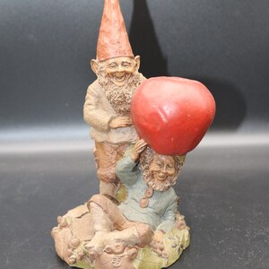 May include: Two gnomes, one with a red hat and the other with a blue shirt, are holding a large red apple. The gnome with the red hat is standing and the gnome with the blue shirt is sitting. The gnomes are made of a brown material and have painted details.