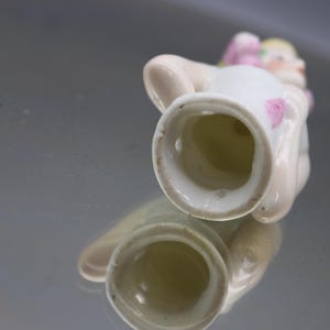May include: A small, white porcelain figurine of a seated figure with a pink bow and accents. The figurine has a detailed face and is reflected in the shiny surface below. The figurine is likely a decorative item.