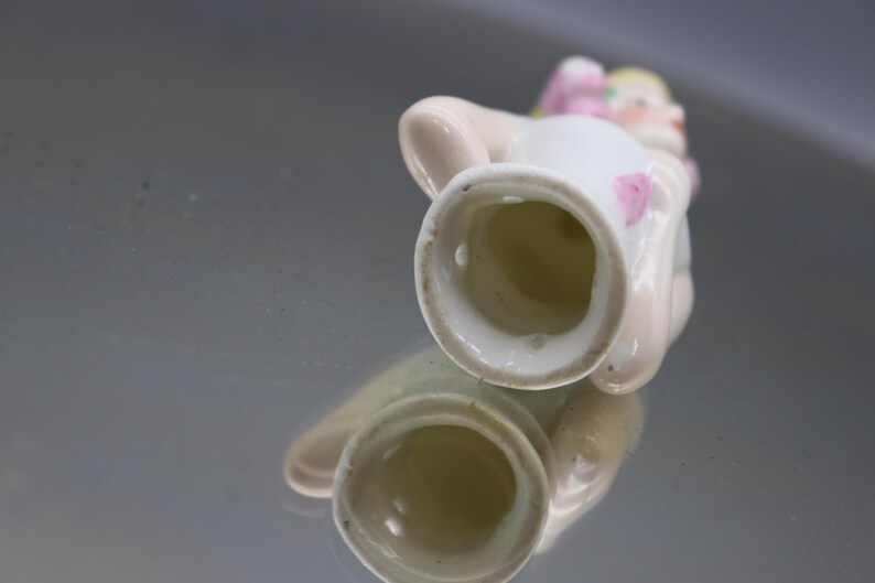 May include: A small, white porcelain figurine of a seated figure with a pink bow and accents. The figurine has a detailed face and is reflected in the shiny surface below. The figurine is likely a decorative item.