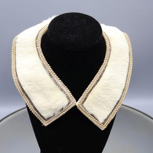 May include: Cream-colored faux fur collar with a pearl beaded trim. The collar is displayed on a black mannequin head. The collar has a soft, fluffy texture and a classic design, suitable for vintage-inspired fashion.